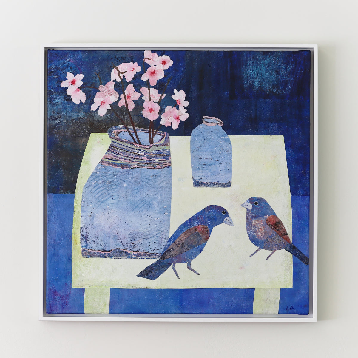 A framed square collage painting featuring a stylized still life with blue birds, pink blossoms, and blue vases on a cream table against a deep blue background, shown against a light neutral wall.