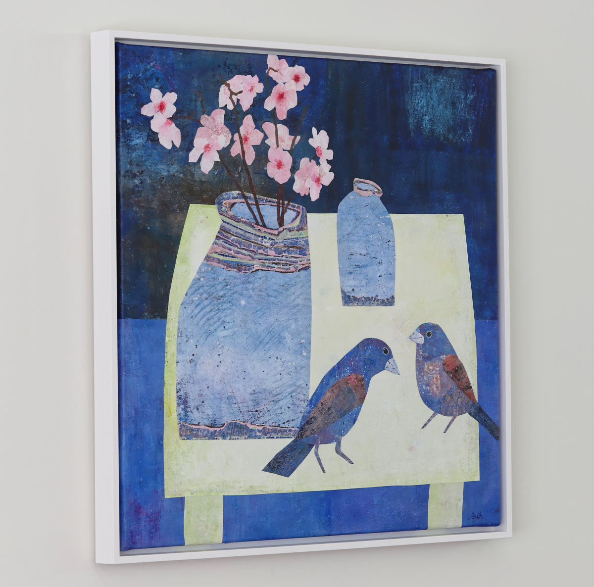 A framed abstract still life collage featuring blue birds, a blue vase with pink blossoms, and a blue bottle on a pale yellow table against a dark blue background, hung on a light gray wall.