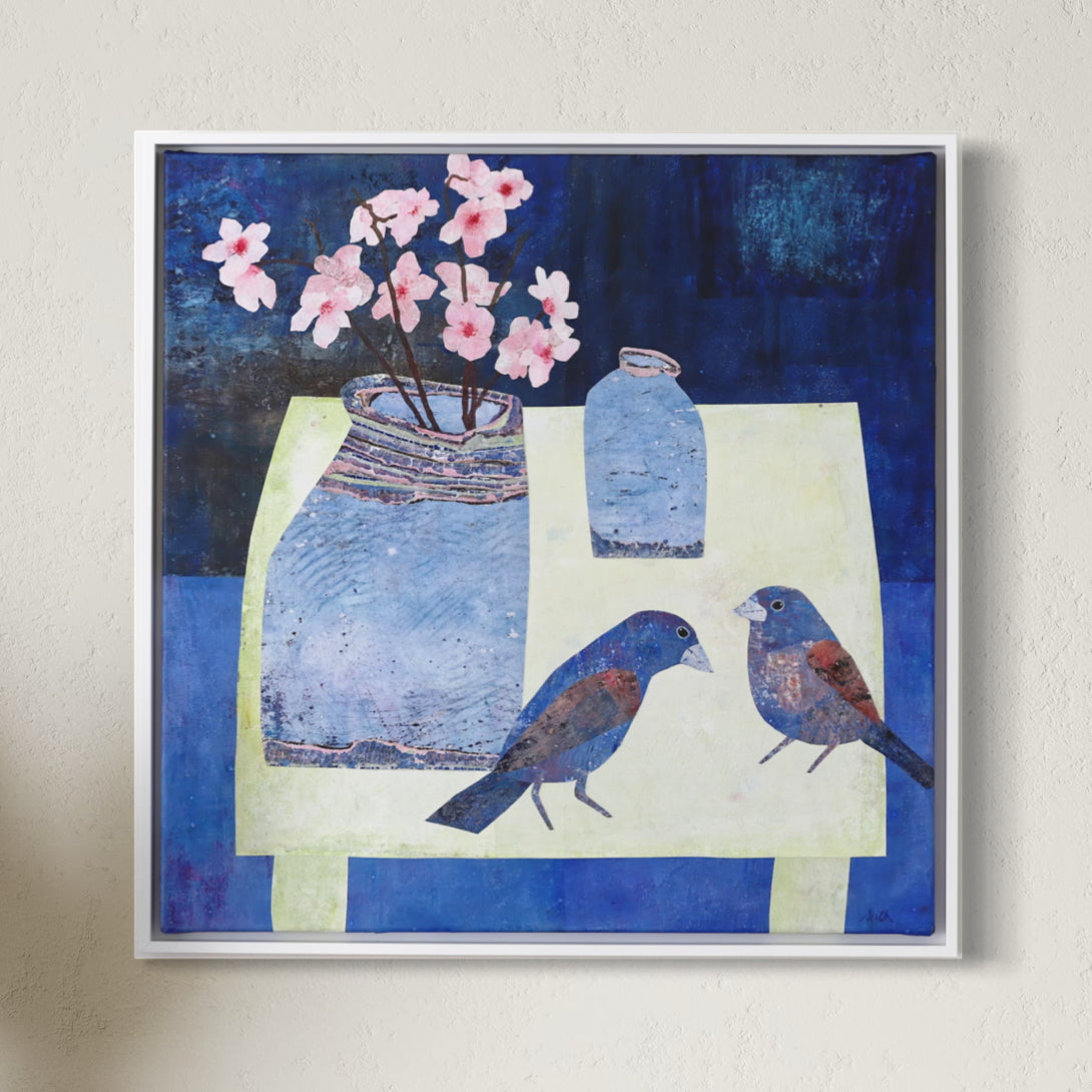 A framed square collage depicting a still life scene with two blue birds, a blue vase with pink flowers, and a smaller blue bottle on a pale yellow table against a dark blue background, hung on a light beige wall.