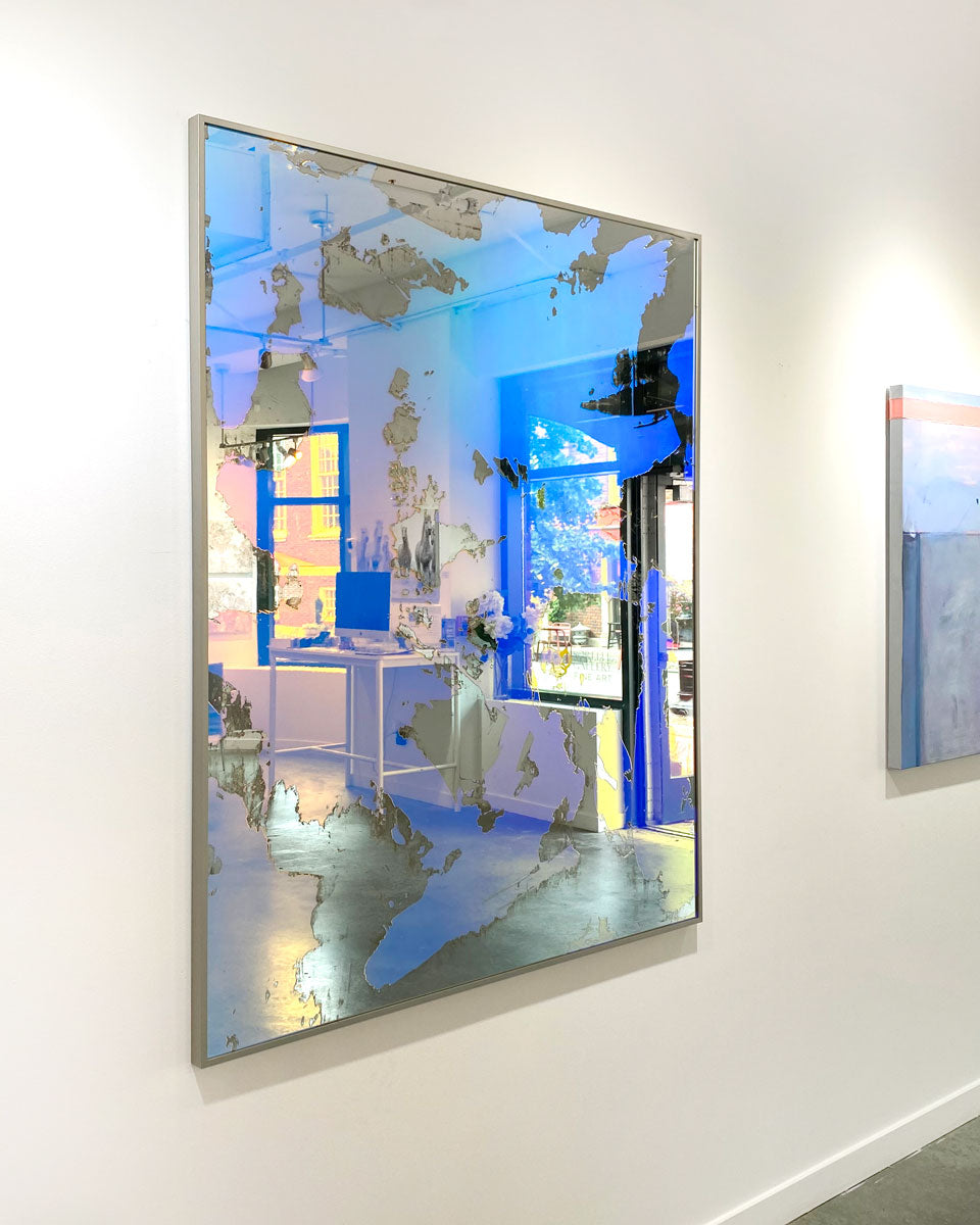 A pink and blue fine art mirror hangs next to a painting on a white wall in an art gallery. 