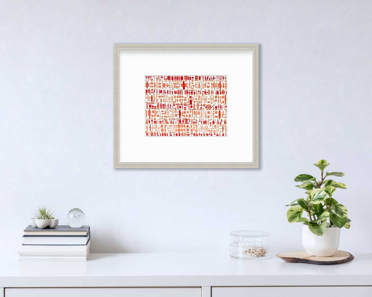A red, white and orange abstract print in a silver frame with a mat hangs on a white wall above a white surface with a stack of books, a round clear container with gold accessories and a potted plant.