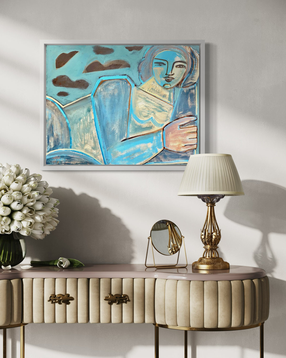 A blue, brown, and yellow painting of an abstracted woman framed in a white floater hanging on a wall over a womans dressing table with a lamp, mirror and vase of flowers.