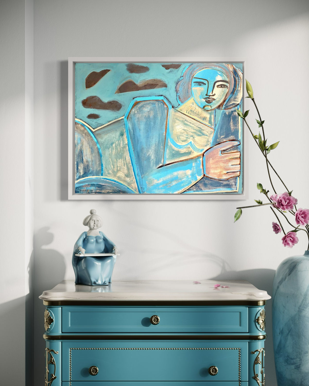 A blue, brown, and yellow painting of an abstracted woman framed in a white floater hanging on a wall over a teal ornate dresser with whiter marble top. A teal woman buddha sits on top and pink flowers lean into view.