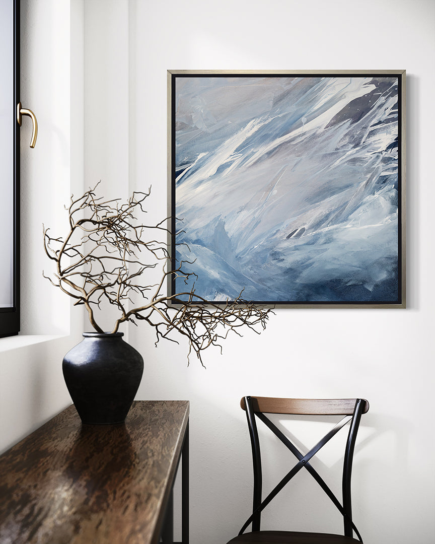 A blue, grey, white and charcoal abstract print in a silver floater frame hangs on a white wall above a wooden chair.  A window is to the left with a wooden table under it and a ceramic pot with twigs sits on the table. 