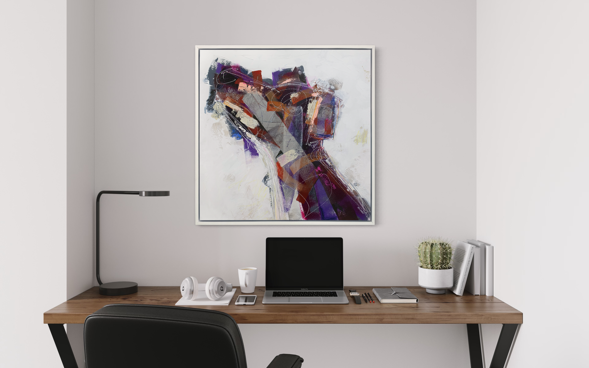 A white, purple, grey and dark orange abstract dog print in a white floater frame hangs on a white wall above a wood desk with a laptop computer, a stack of books, a pair of headphones and a modern black desk lamp.