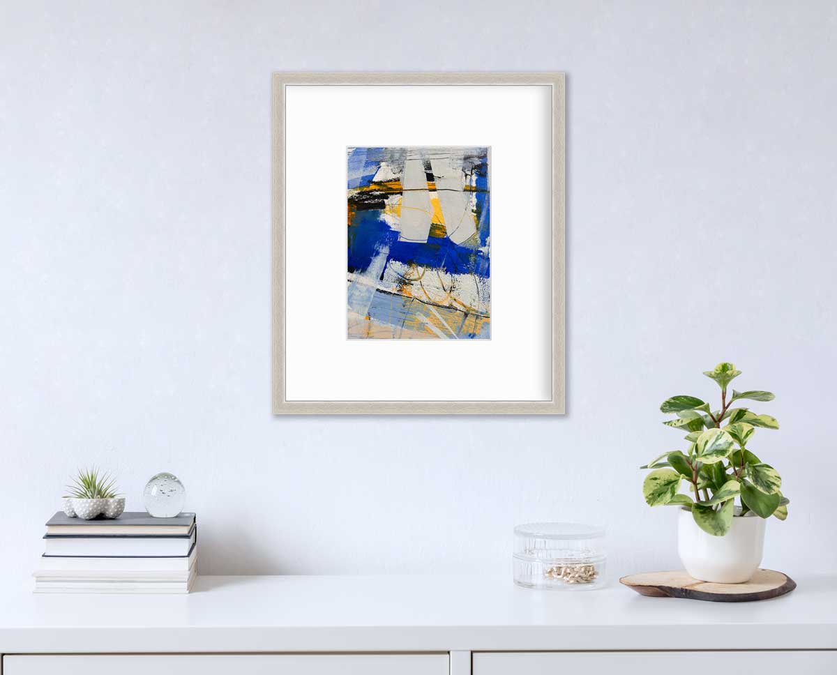 A blue, grey and yellow abstract print in a silver frame with a mat hangs on a white wall above a white surface with a stack of books, a round clear container with gold accessories and a potted plant.