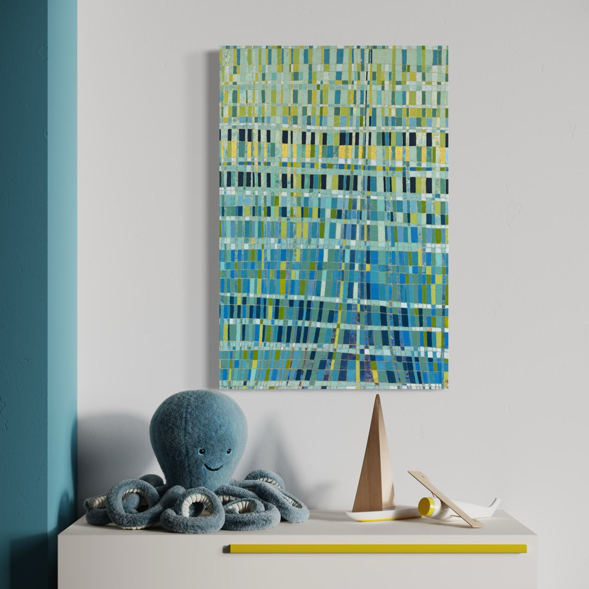 An abstract painting featuring a textured pattern of blue, green, and yellow rectangle, displayed on a light gray wall above a white side table with a blue octopus plush toy and a wooden sailboat decoration.