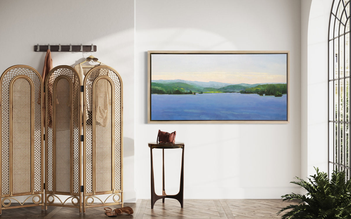 A blue landscape art print framed in a white oak frame hangs on a white wall between a large arched window and a wall rack with sweaters hanging from it and a free-standing wicker screen. 