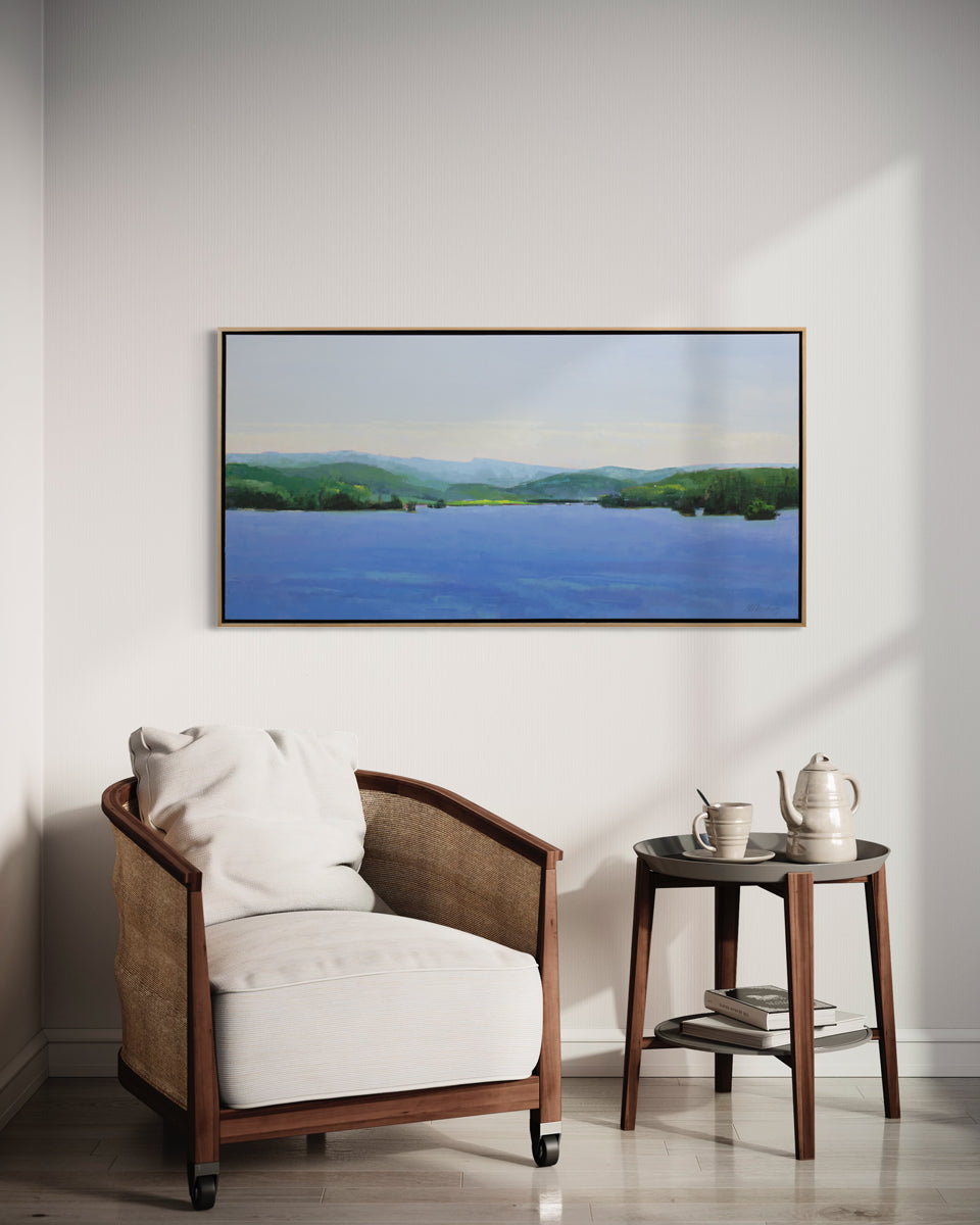A blue and green landscape painting framed in a light wood frame hangs on a light wall above a brown armchair and a contemporary wood end table with books stacked and a teapot and teacup. 
