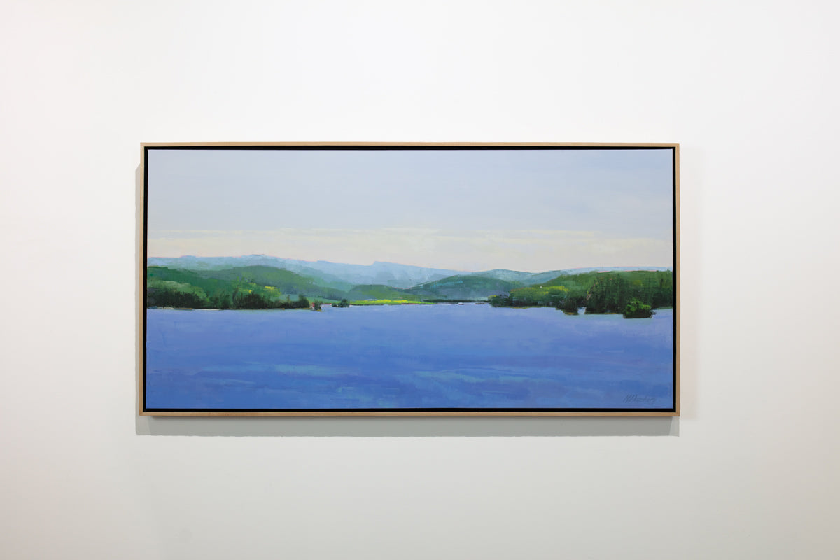 A blue and green landscape painting framed in a light wood frame hangs on a white wall. 