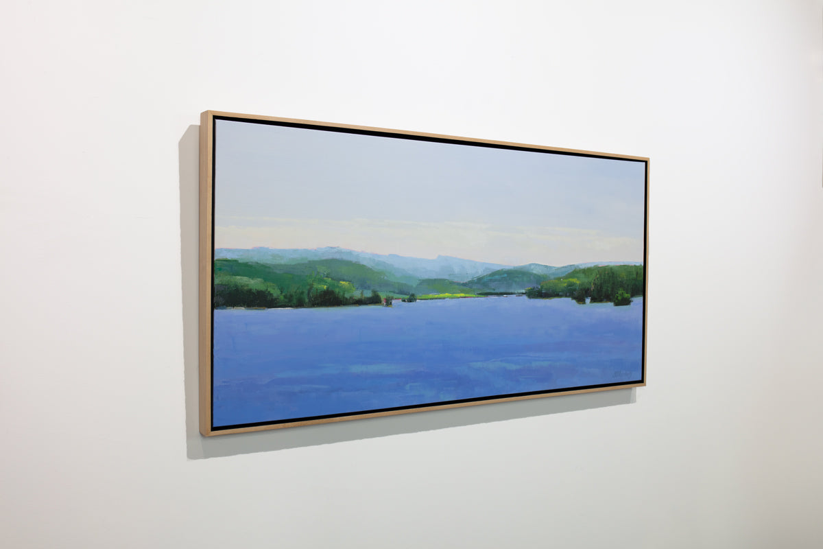 A blue and green landscape painting framed in a light wood frame hangs on a white wall.