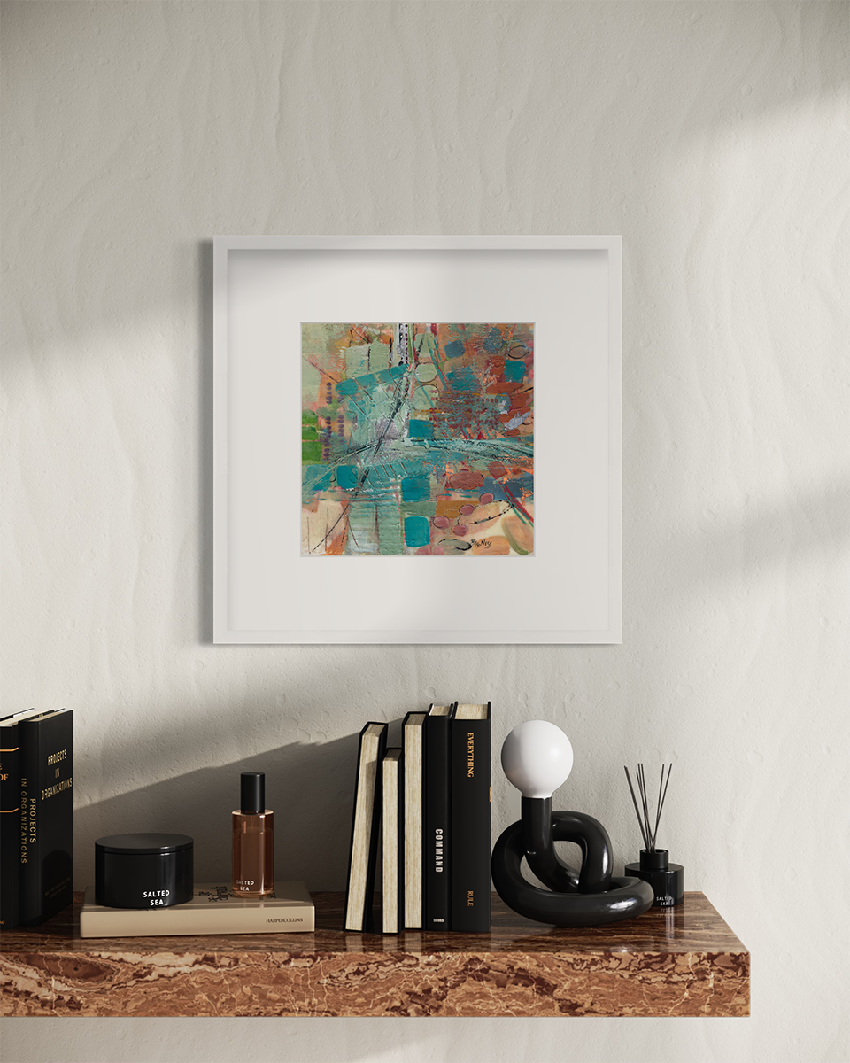 An abstract multicolored print, matted and framed in a white frame hanging on a white wall above a brown marble shelf with books, a small modern light and candles.
