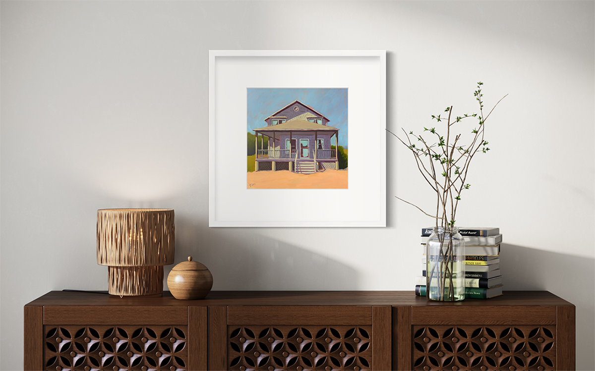 A blue, lavender and peach contemporary beach house print in a white frame with a mat hangs on a white wall above a dark wood credenza with a stack of books, a clear jar with stems and a woven lamp.
