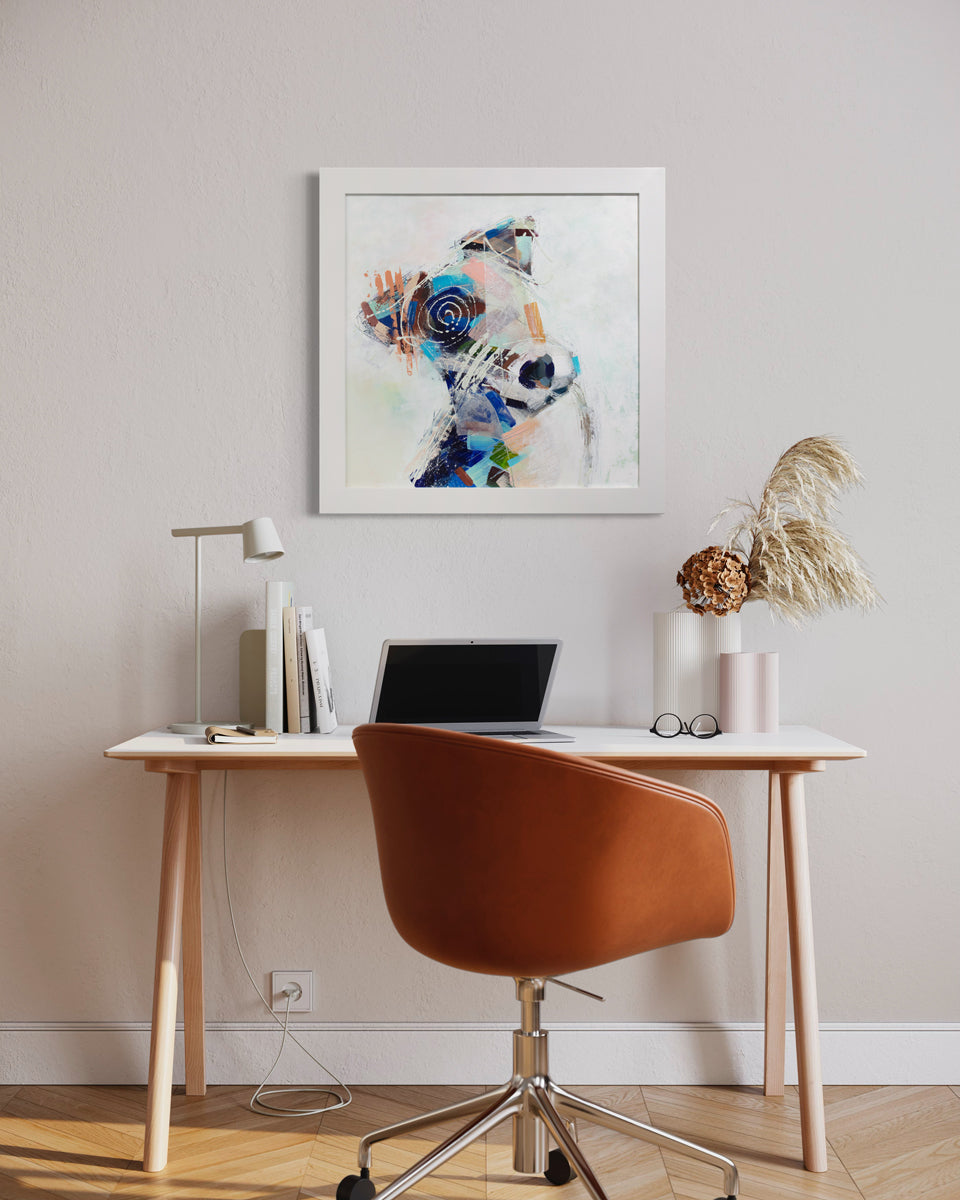 An abstracted painting of a dog in a white frame hangs on a light wall above a modern wood desk and chair with decor. 