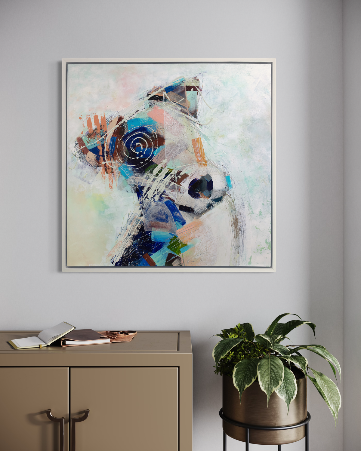 A multicolored abstract dog print in a white floater frame hangs on a white wall above a beige metal cabinet and a potted plant in a riser.