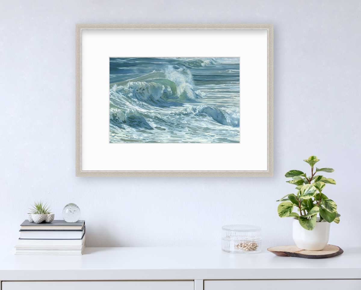 A blue, teal, white and light green abstract water wave print in a silver frame with a mat hangs on a white wall above a white countertop with a stack of books, a clear round container with gold accessories and a potted plant.