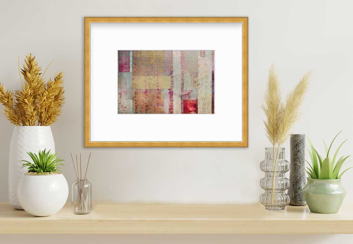 A red, yellow and grey abstract print in a gold frame with a mat hangs on a white wall above a wood surface with several vases with wheat stems and greenery.