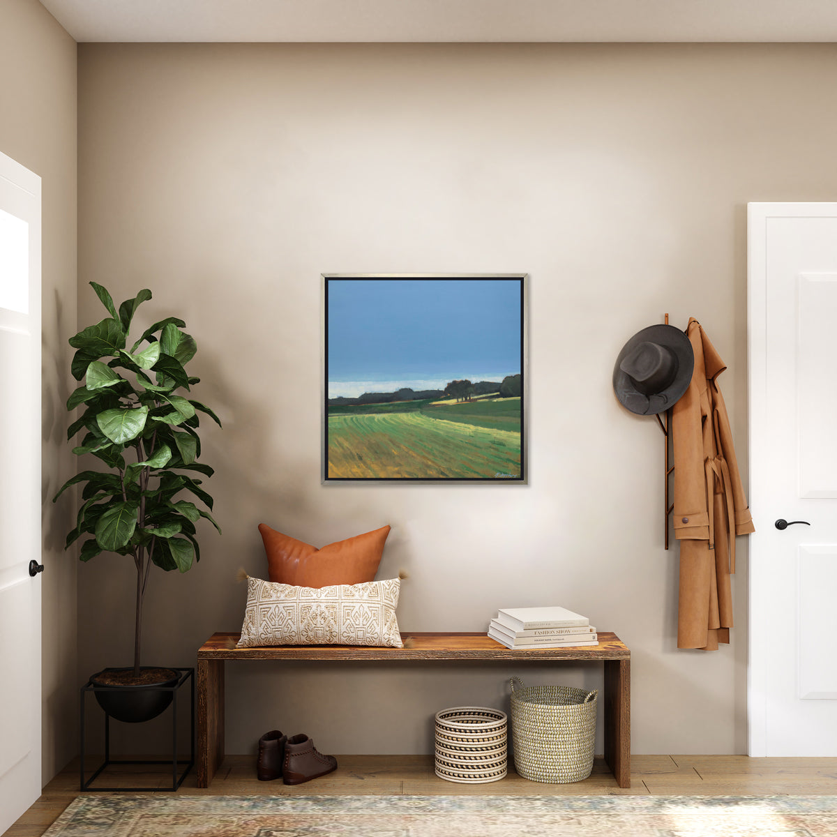 A landscape print in a silver floater frame hangs on a beige wall above a live edge wood bench with pillows, a stack of books, two woven baskets and a pair of shoes.