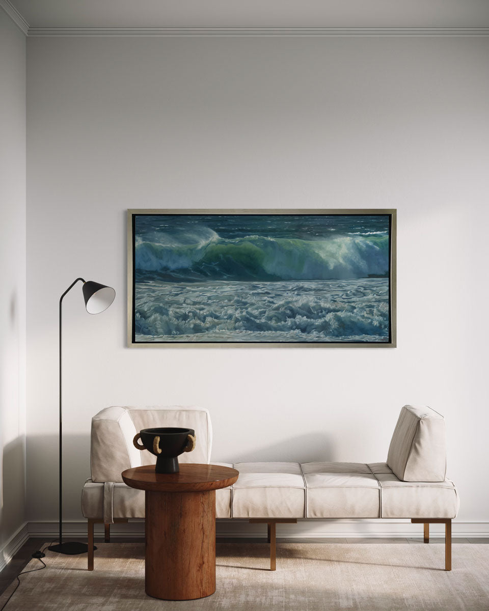 A painting of blue-green waves with white foam hangs in a silver frame on a white wall with a padded bench, small side table and standing lamp in front of it.