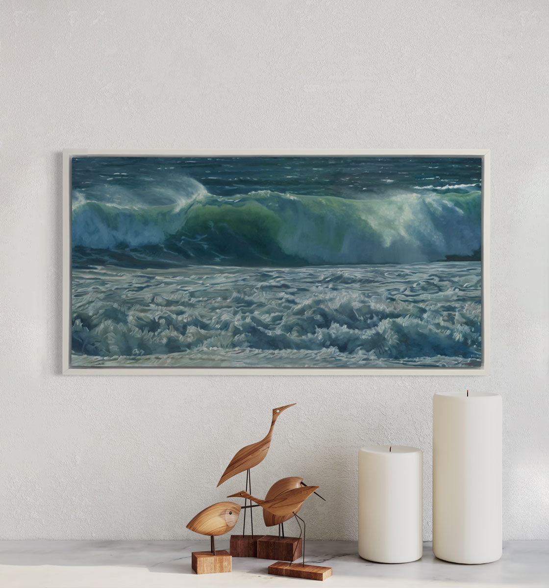 A painting of blue-green waves with white foam hangs in a white frame over a mantle with a bird sculpture and candles below it.