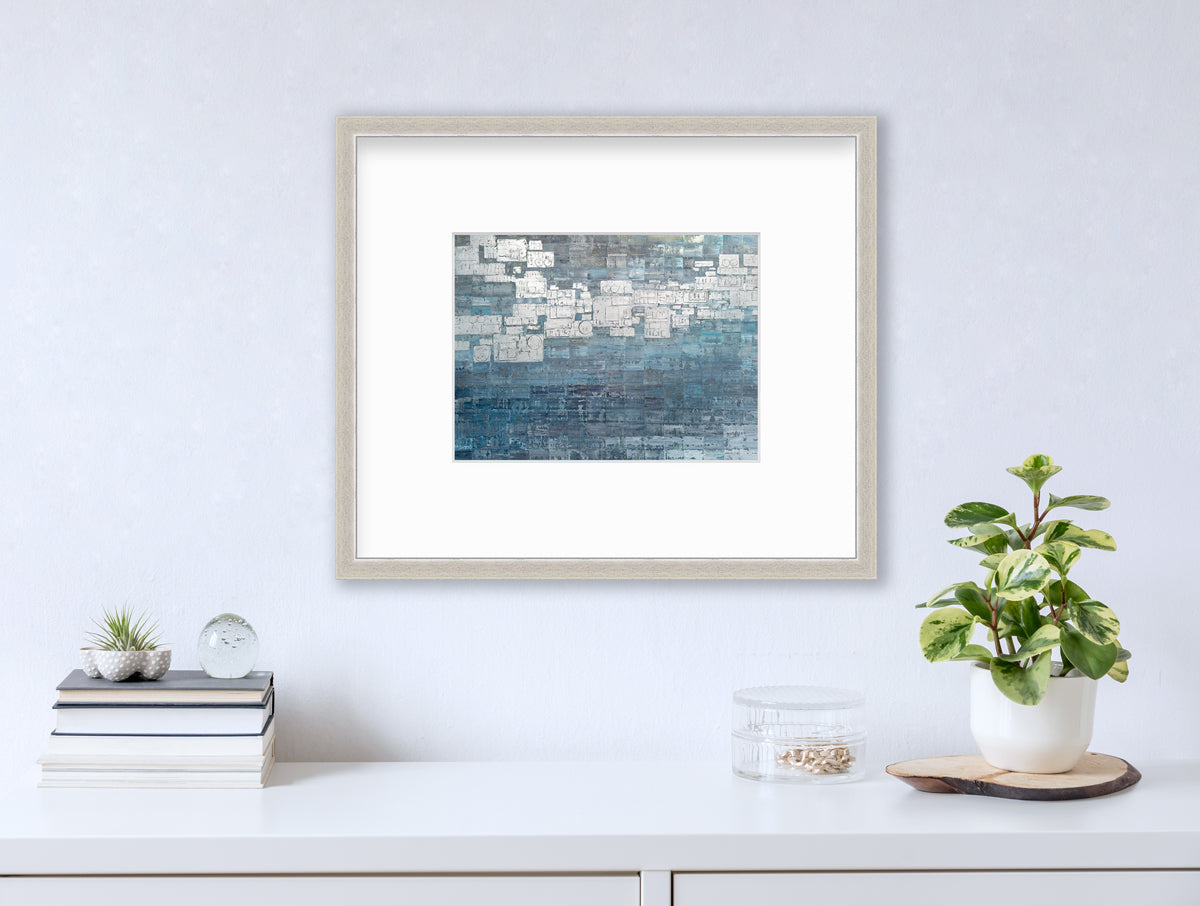 A blue, white and grey abstract print in a silver frame with a mat hangs on a white wall above a white surface with stack of books, a round clear container with gold accessories and a potted plant.