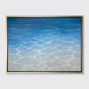 A tonal blue, beige and white seascape print in a silver floater frame hangs on a white wall.