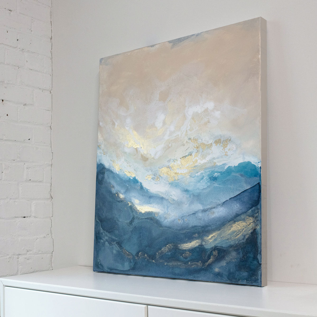 An abstract blue and gold painting rests against a light wall on top of a white dresser. 