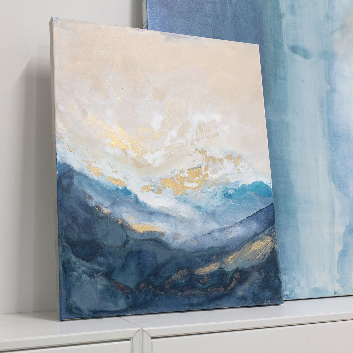 A blue and gold painting rests against an abstract painting behind it on top of a white dresser. 