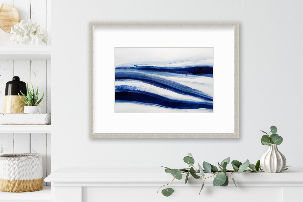A dark blue and white abstract print in a silver frame with a mat hangs on a white wall above a floating white shelf, a white vase with eucalyptus leaves and a built in bookshelf.