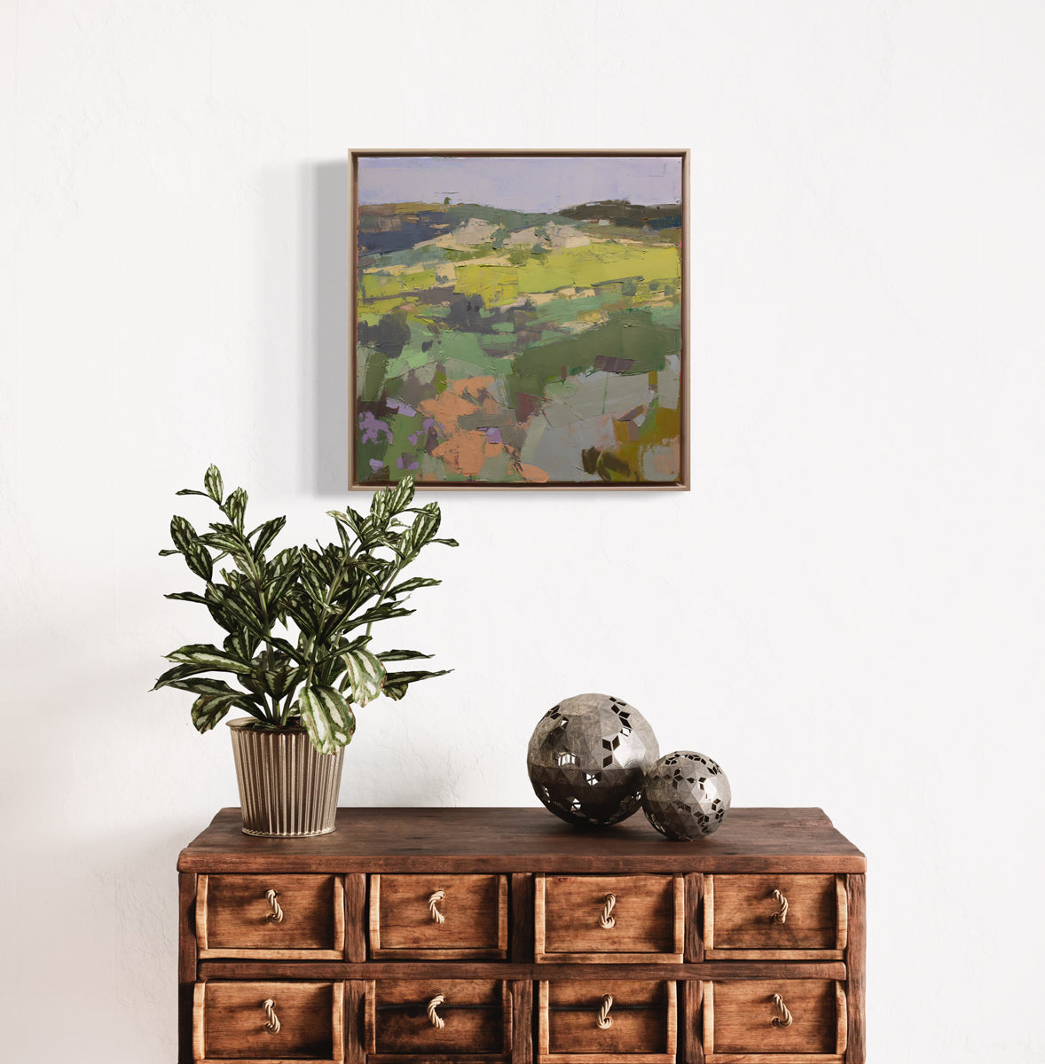 A textured abstract landscape painting hangs on a white wall above a wooden storage table with a potted plant and modern rustic table decor. 
