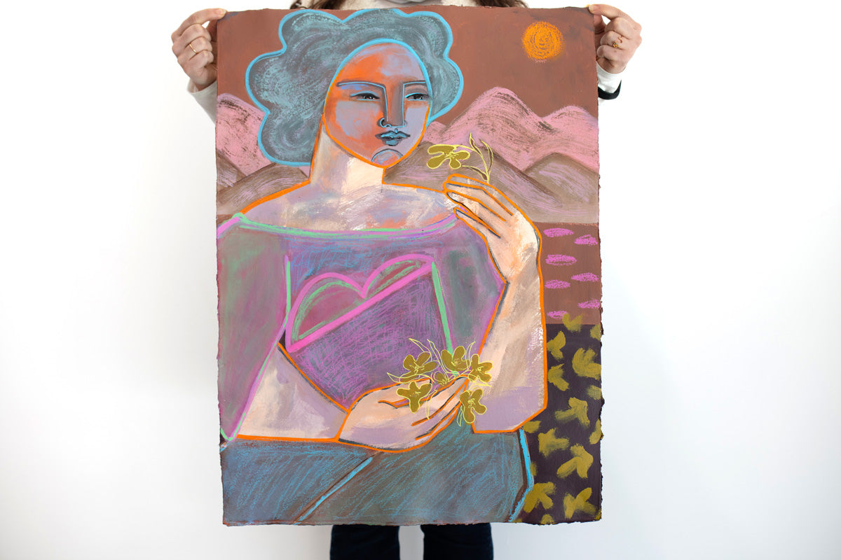 A woman holds a warm-toned, abstract painting on paper of a woman in front of a mountainscape in front of a white wall. 