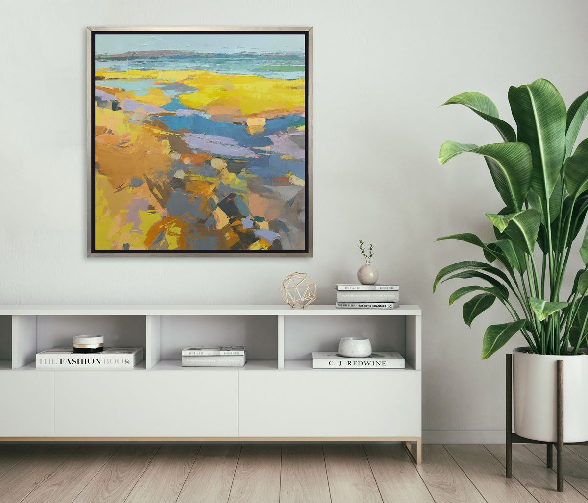 A yellow abstracted landscape print framed in a silver frame hangs on a white wall above a modern white table with decor and a large potted floor plant. 
