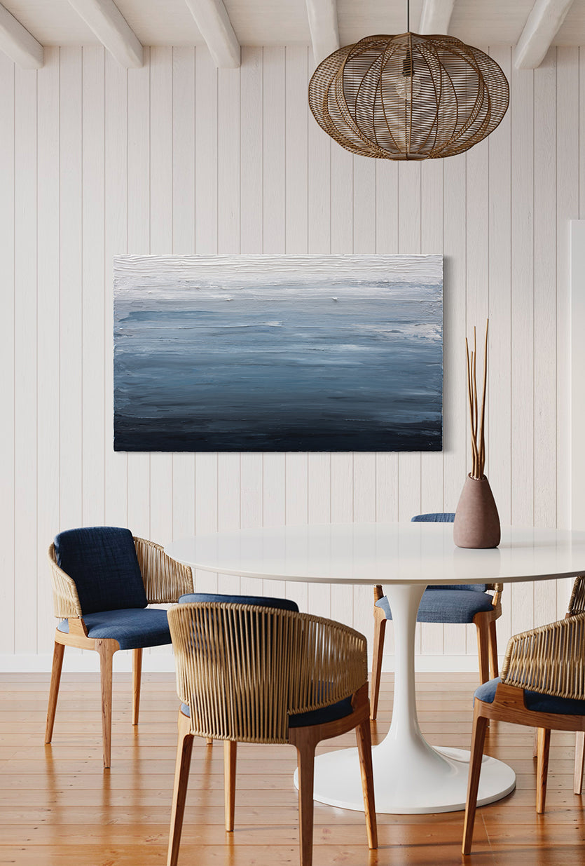 A thickly painted painting in dark navy blue, light blue and white by Teodora Guererra hang on a white wall in a kitchen. A white pedestal table surrounded by modern wicker type chairs with blue cushions sits in front of the paintings with a ceramic vase and sticks sitting on the table. Over the table hangs a wicker light and a door to the right can be seen in apple green.