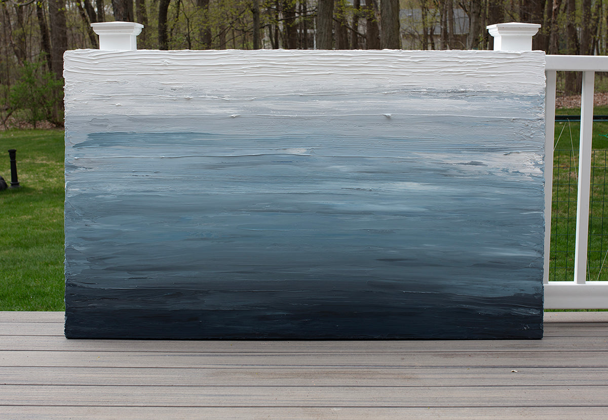 A dark navy blue, light blue, blue, and white thickly textured abstract painting sitting on a deck outside in natural light by Teodora Guererra.