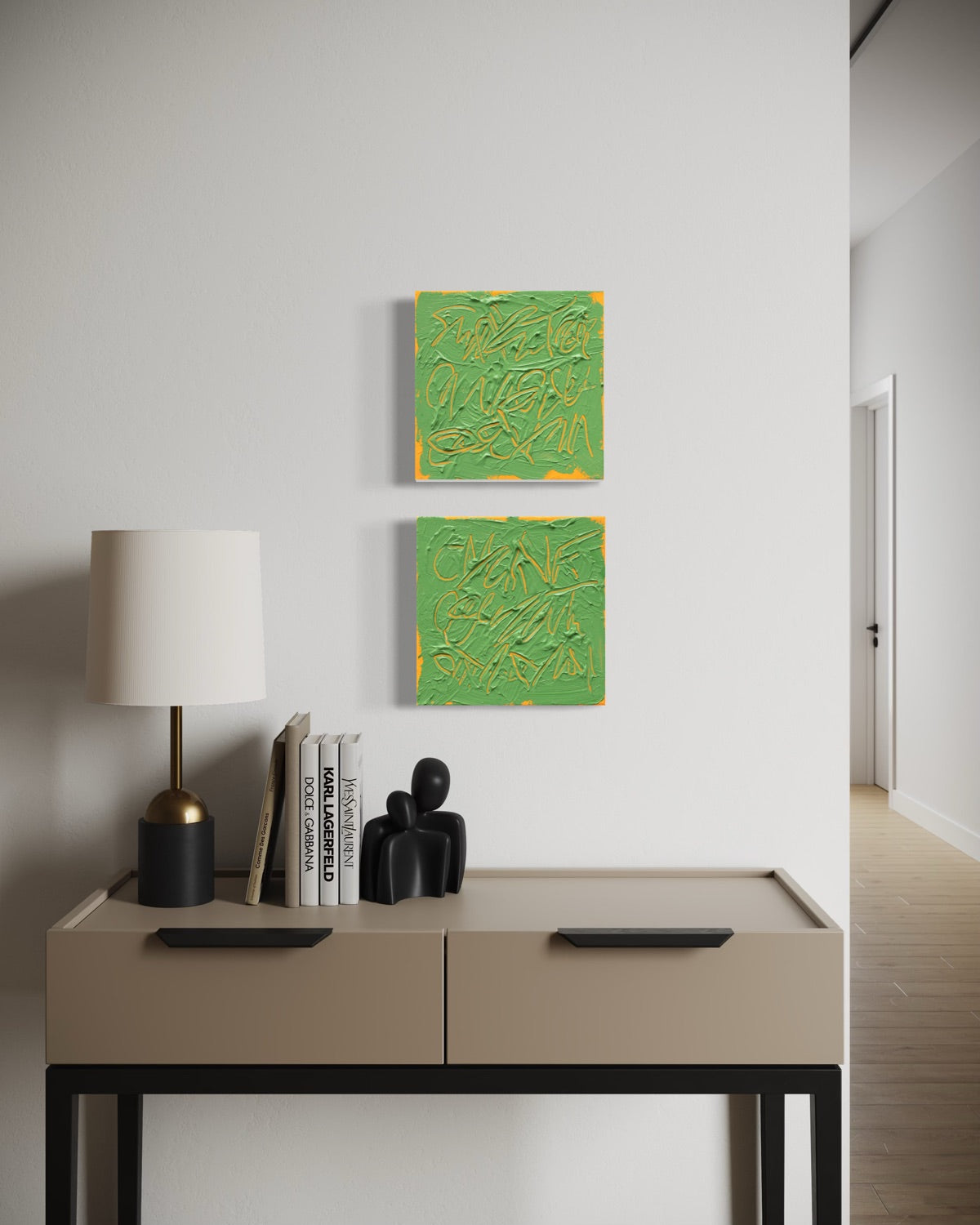 Two square abstract text based paintings with thick green paint and tangerine gestural lines, hanging on a white wall above a beige console table with a lamp and books.