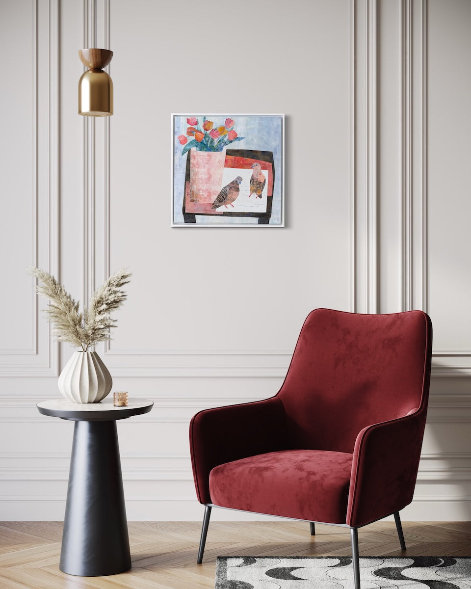 A modern red velvet armchair in a styled living room setting, positioned next to a black side table with a vase of dried grass and a framed floral collage on a light gray paneled wall.