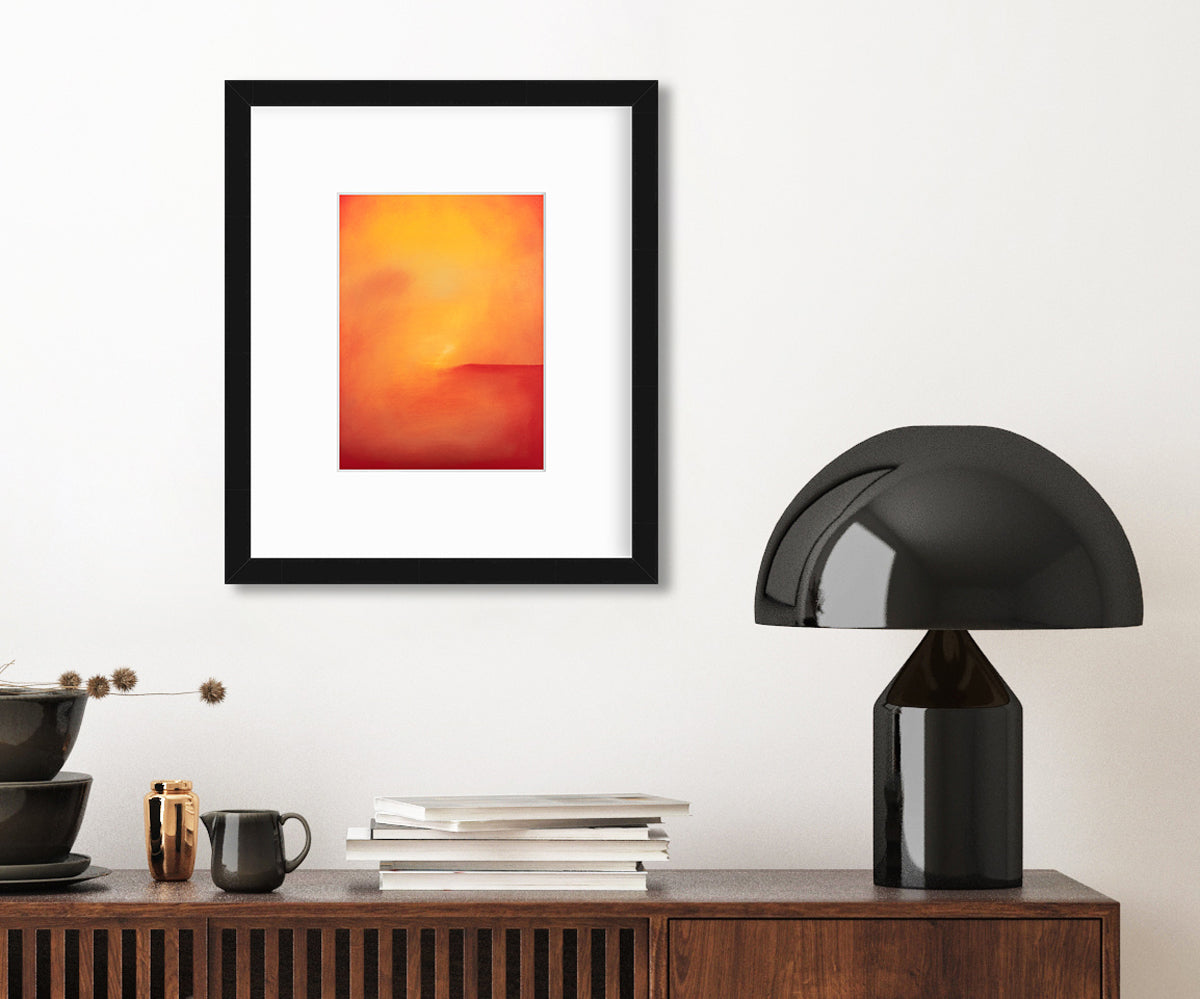 An orange and red abstract print in a black frame with a mat hangs on a white wall above a dark wood credenza with a stack of brown bowls, a stack of books and a modern black table lamp.