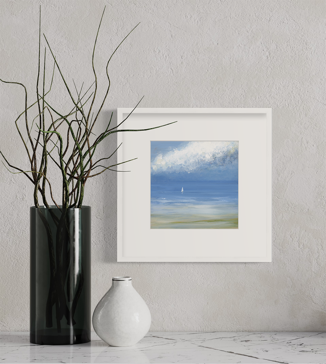 A blue, light green and white abstract seascape print with a small sailboat in a white frame with a mat hangs on a white wall above a marble surface with a small white vase and a cylindrical vase with stems.