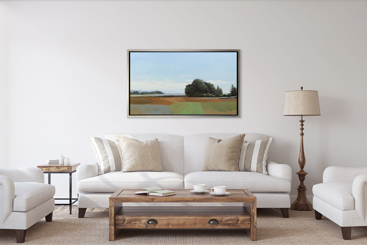 A landscape print in a silver floater frame hangs on a white wall above a white sofa with neutral pillows, a dark wood floor lamp and a stained wood coffee table.