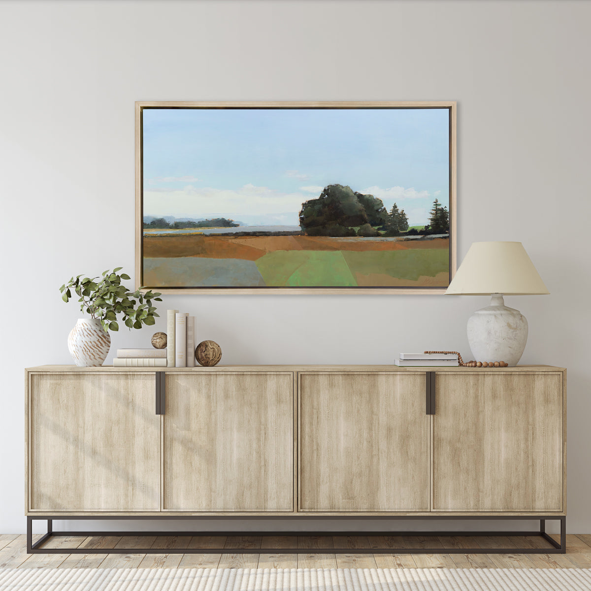 A landscape print in a white oak wood floater frame hangs on a white wall above a natural wood credenza with a white table lamp, a stack of books and a white vase with eucalyptus stems.