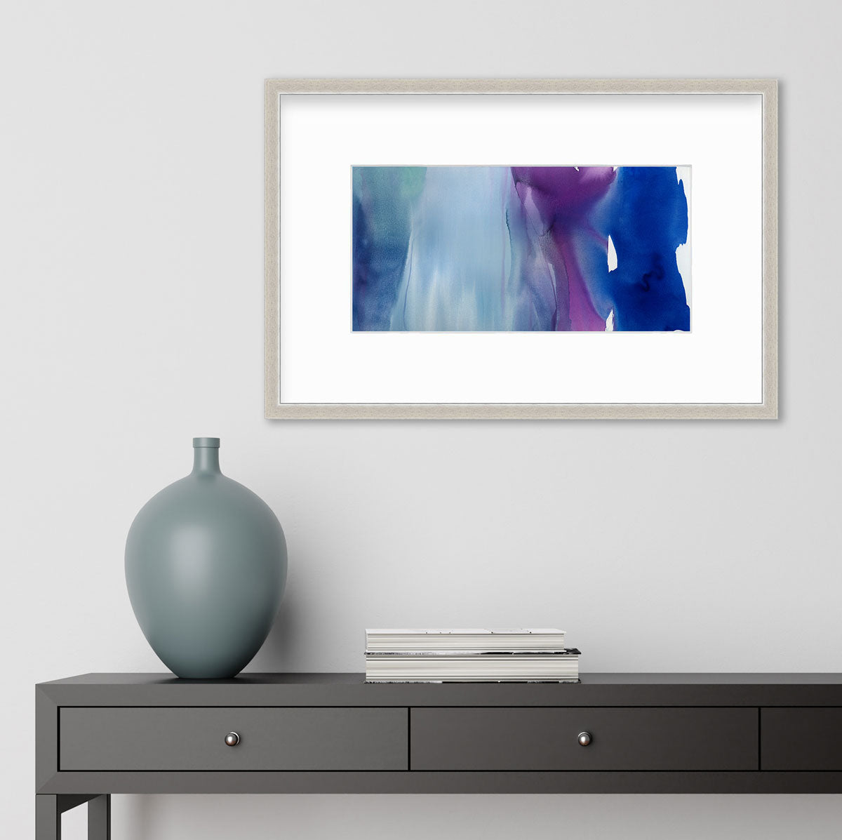 A blue, purple and white abstract print in a silver frame with a mat hangs on a white wall above a black desk with books and a muted teal vase.