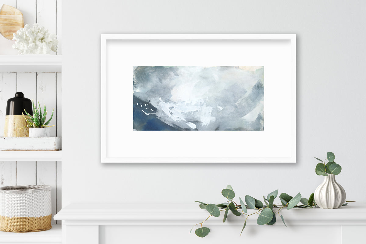A blue, grey and white abstract print in a white frame with a mat hangs on a white wall above a white shelf with eucalyptus coming out of a small vase and a built in shelf with accessories.