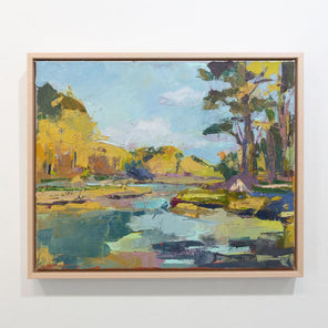 A blue and yellow abstract landscape painting framed in a natural floater frame hangs on a white wall.