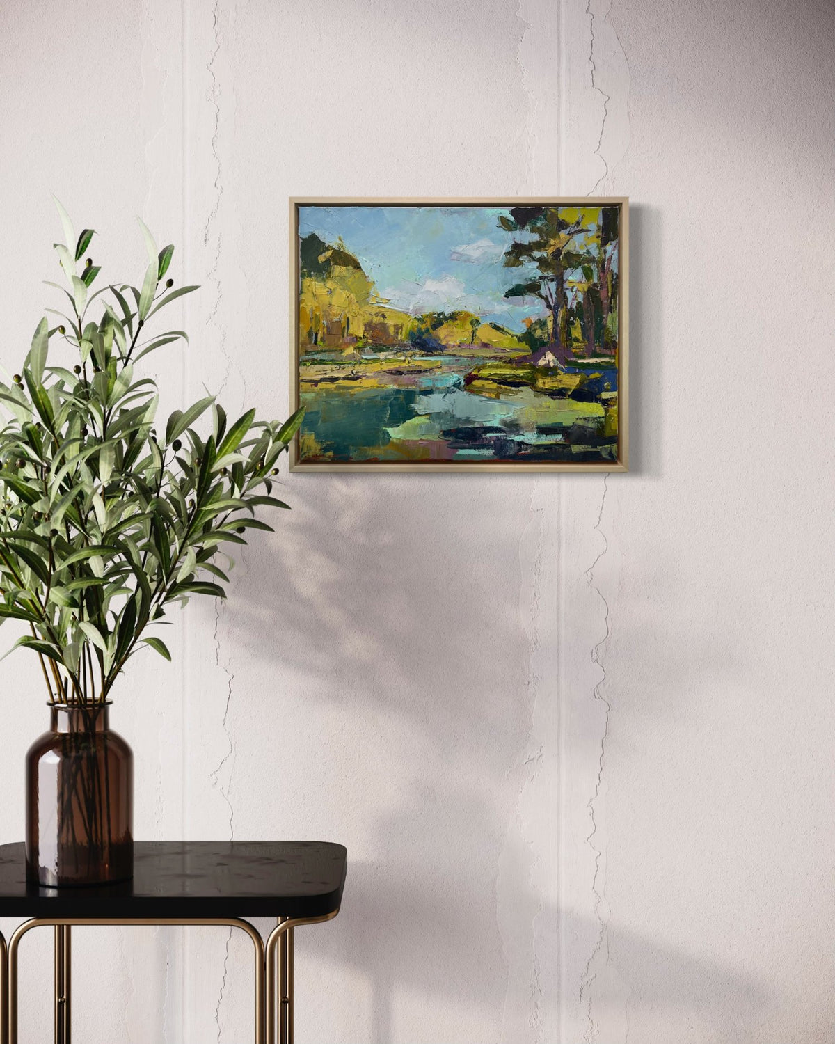 A blue and yellow abstract landscape painting hangs on a white wall with a table to the left with a vase holding eucalyptus branches.