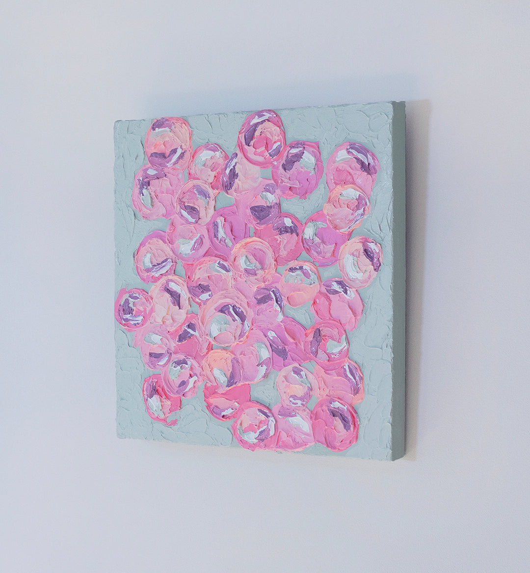 A very textured abstract painting with celadon background and circle type abstract flowers in pink, peach, plum and white hangs on a white wall, side view.