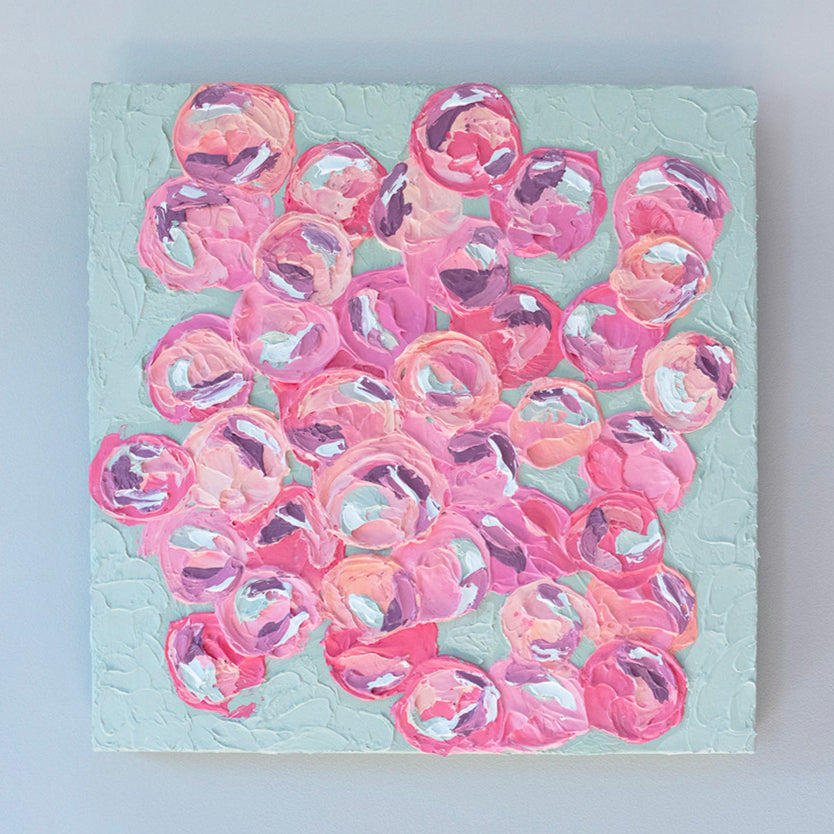 A very textured abstract painting with celadon background and circle type abstract flowers in pink, peach, plum and white hangs on a grey wall.