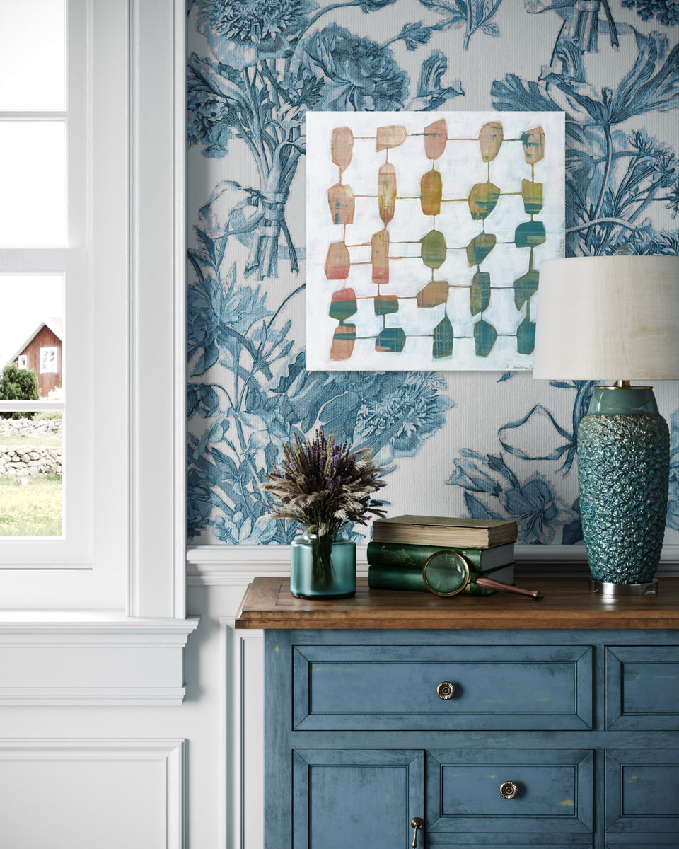 An abstract painting hangs on a wallpapered wall above a blue dresser with teal table decor. 