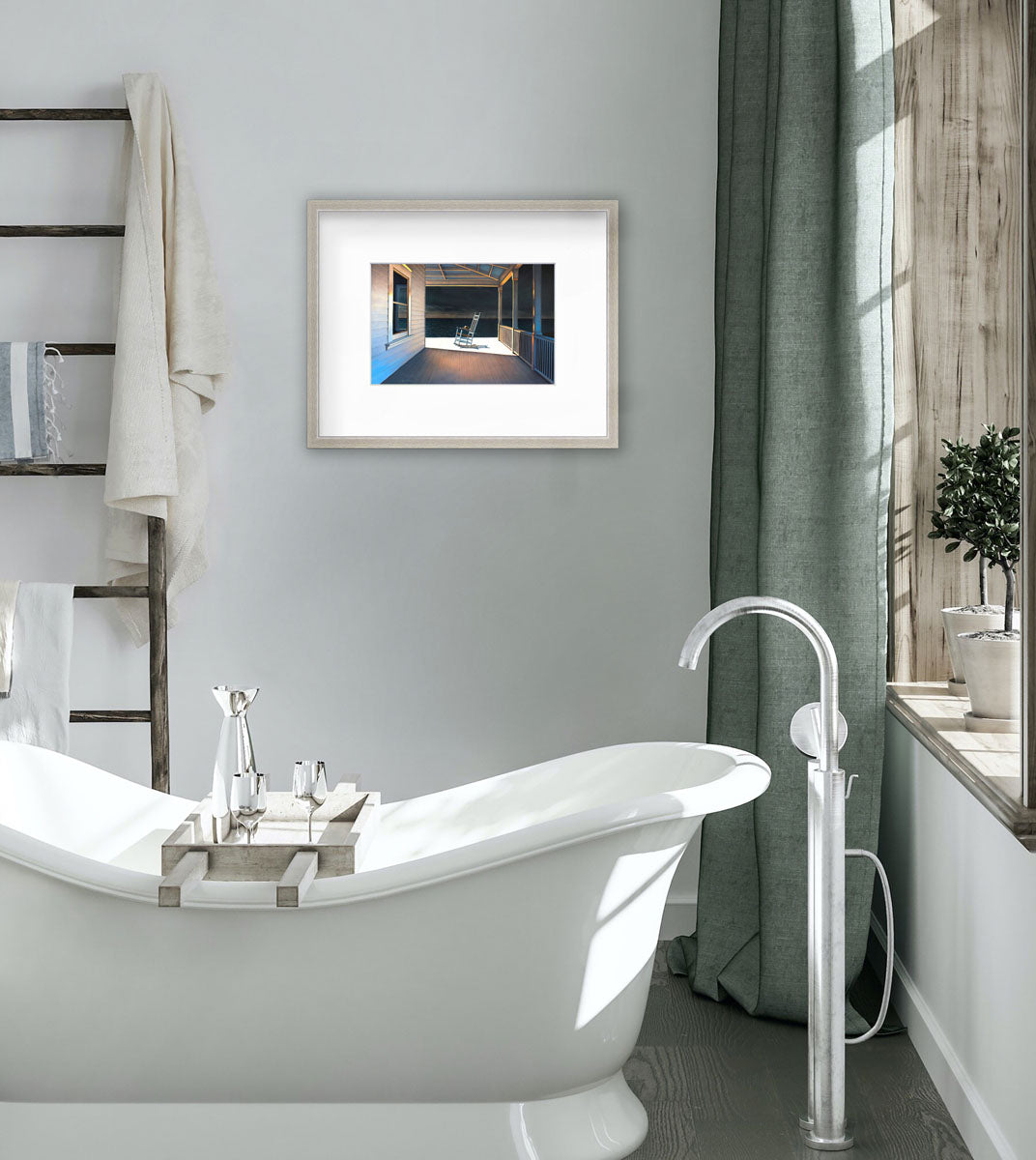 A print of a white rocking chair on a deck of a house with a high contrast light on the chair matted in silver frame hangs in a bathroom. the print hangs to the left of a large window and above a soaking tub.