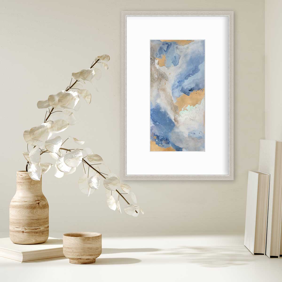A blue, teal, beige and gold abstract print in a silver frame with a mat hangs on a white wall above a stack of books and a wood vase with white leaf stems.