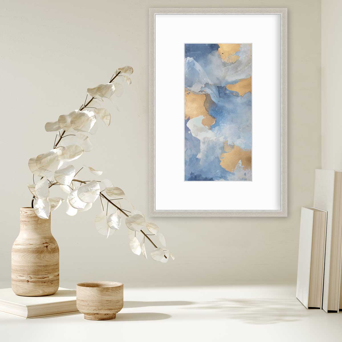 A blue, teal, beige and gold abstract print in a silver frame with a mat hangs on a white wall above a stack of books and a wooden vase with white leaf stems.